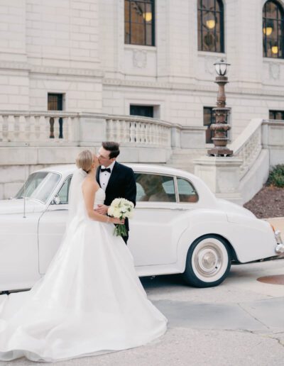 Groom wearing a custom wedding tuxedo from Tailored Gents at Savile Row kissing his bride in front of a car.
